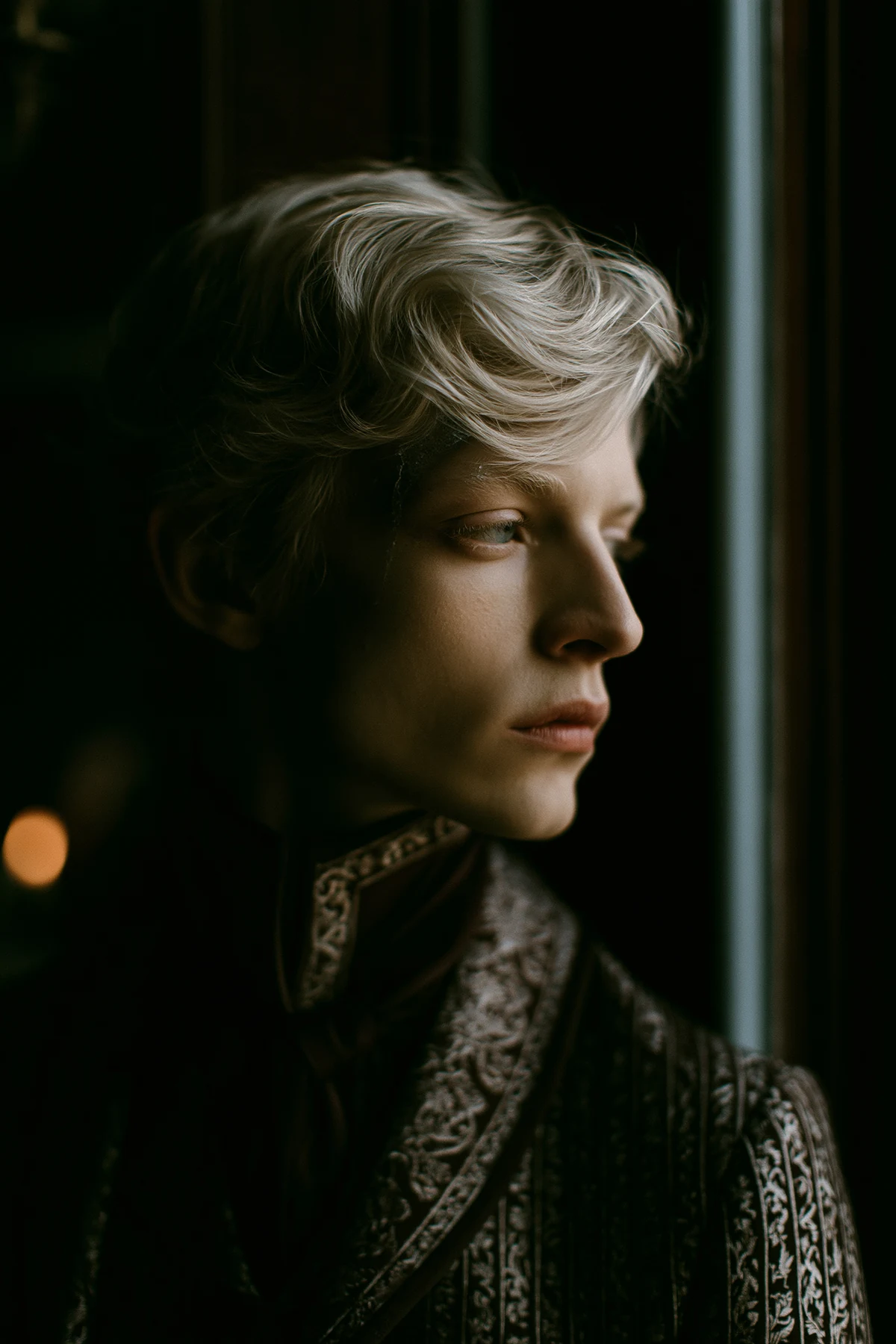 A blond figure in an ornate coat beside a window.